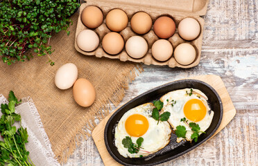 Top view of raw hen eggs in egg box and fried eggs in a frying pan on wooden background with burlap and greens