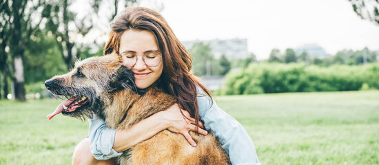 Women hugging dog in the summer park. Cheerful lady with long dark hair in blue jacket hugs and strokes friendly old dog sitting on lush green meadow of public garden on nice day. - Powered by Adobe