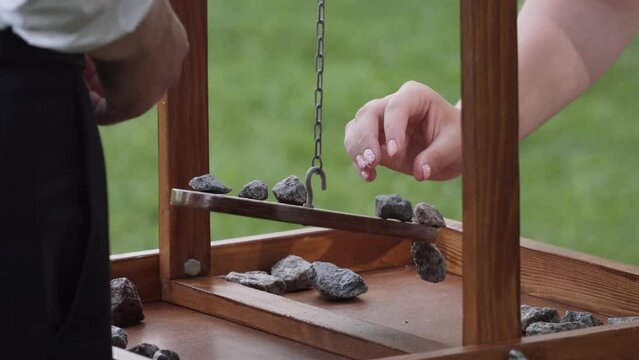 Wooden disc of game balancer is hung by hook on chain, swinging over board on green lawn. Woman puts stone on board for balance. Game chips fall, game over. Man takes the glasses.