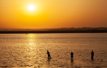 Long shot of a sunset at the beach with silhouette of three fishermans