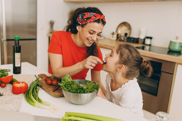 Happy mom and daughter at kitchen, cooking together, mother giving child cherry tomato to try, woman wearing red t-shirt and stylish bandana, girl dressed in white, vegetables and utensils on table