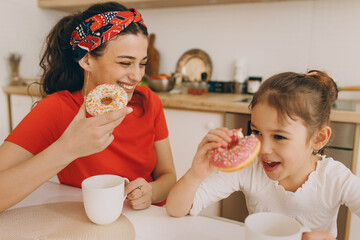 Stylish attractive mother in red cooked tasty doughnuts along with her daughter, drinking tea and...