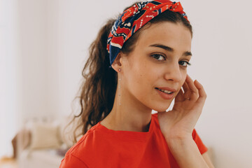 Horizontal indoor portrait of young charming mysterious female of 20s in red colorful bandana over forehead, hair tied in ponytail, touching face gently while looking at camera, dressed in t-shirt
