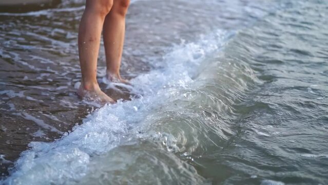 female legs and feet walking along sea water waves on sandy beach. wounded legs woman walks at seaside surf. Splashes of water and foam in slow motion. Girl in ocean at sunset or sunrise.