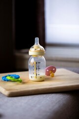 A portrait of a baby bottle with a bit of formula still in it standing on a wooden plank. The glass nursing bottle still has some milk in it and has a rubber dummy nipple, or bottle teat.