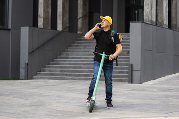 Delivery man using smartphone and riding push scooter in the city. Young delivery man with thermal backpack using smartphone while riding scooter on the background of modern buildings.