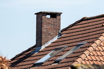 A portrait of a chimney on a roof of a house. The roof has ceramic tiles and two skylight windows...