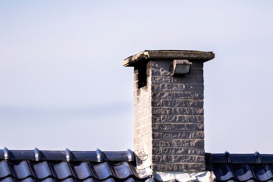 A portrait of a brick chimney on the top of a roof with blue ceramic tiles on a sunny day.