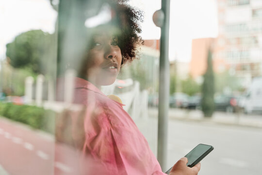 african american woman with bulky curly hair is sitting inside of the outdoor bus stop and using her smartphone