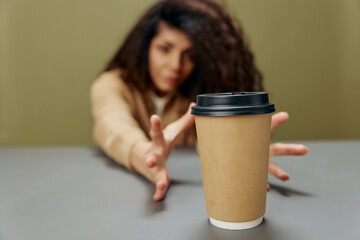 Happy smiling tanned curly Latin female reach for a cup to-go to enjoy coffee sit at the table isolated over olive green background. Copy space Mockup Banner. Coffee lover offer. Wide angle