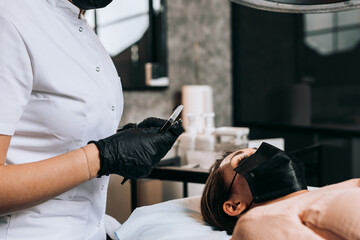 Cosmetologist measures the proportions of the eyebrows with the ruler. Micropigmentation work flow in a beauty salon. They are iwearing face protective masks due to Covid-19 pandemic.