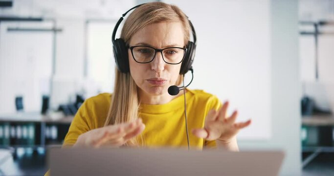 Customer service operator in headset having conversation with clients in bright call center office. Caucasian woman sitting in front of laptop and answering calls.