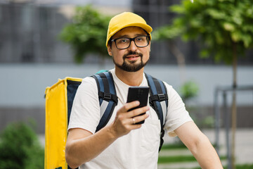 Portrait of delivery man with yellow backpack and a scooter looking for the client's address in phone. Courier checking customer address on his phone, food delivery outbreak. Fast delivery.