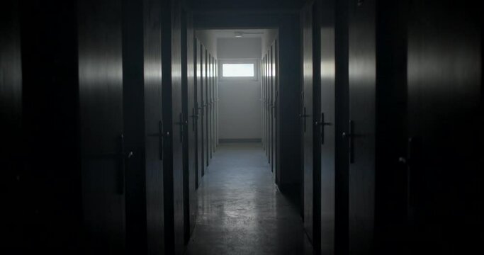 A dark prison room with a long corridor. Lots of cell doors for detainees. The beam of light is moving. At the end of the corridor there is a bright spot of a window.