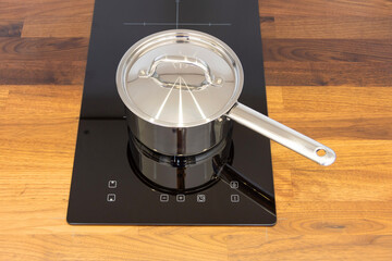 Saucepan ladle with lid on an induction hob built into a wooden kitchen worktop, top view.