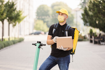 Obraz premium Handsome young man uses an electronic scooter to deliver goods in a cardboard box from online store. Concept of delivery of products in individual packages. Courier in a mask and gloves.
