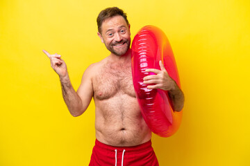 Middle age caucasian man holding inflatable donut isolated on yellow background pointing finger to the side