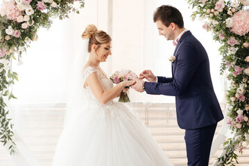 The groom wears a ring to the bride at the ceremony against the backdrop of a round arch of roses