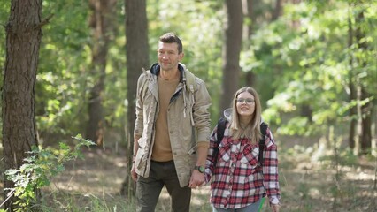 Happy Caucasian man and woman holding hands walking with backpacks in sunny forest outdoors. Relaxed couple enjoying travelling in woodland on sunny day. Age difference and happiness