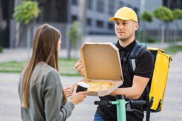 Front view of a delivery man coming to the building of customer and meet young caucasian woman. Girl taking pizza box and cup of coffee from courier. Shipping or delivery service concept.