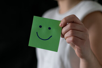 Hand holding green paper cut happy smile face on black background , positive thinking, mental...