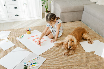 A girl draws hearts for his mother sitting on carpet floor in living room, cocker spaniel dog lying...