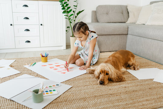 A girl draws hearts for his mother sitting on carpet floor in living room, cocker spaniel dog lying nearby