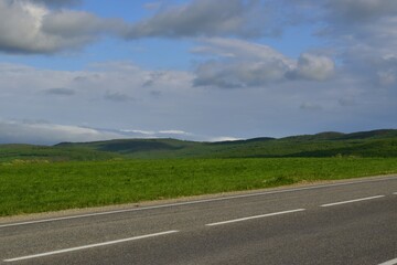The road is along the green hills against the background of blue sky and fluffy clouds. Russia.