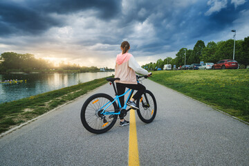 Woman riding a mountain bike on the road near lake at sunset in summer. Colorful landscape with sporty girl riding a bicycle, road, green grass and cloudy sky in park.  Spring. Sport and travel. Cycle