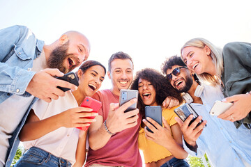 Low angle of Multiracial happy group of friends using phone and smiling together outside. People addicted to technology and social media
