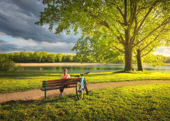 Woman sitting on bench and mountain bike, green trees and lake at sunset in spring. Colorful...