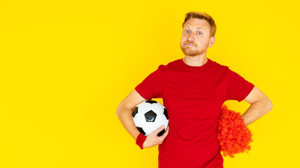 Isolated unhappy soccer supporter man fan in red shirt holding a football ball over yellow...