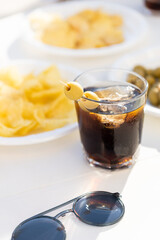 Table full of snacks and a glass of vermuth during a sunny day. Chips, olives and vermut outside with sunglasses on a table.