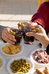 Three people clinking with vermouth glasses in a sunny day. Just three arms cropped above a table full of snacks. Friends enjoying drinks and food.