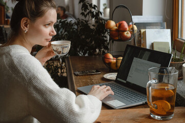 Young freelancer working on laptop at home. Woman using computer on the kitchen for work. Distant job, working remotely in cozy vintage house. Searching education on-line. Adult studding by window