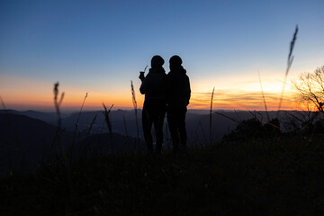 Silhouette of a couple with a yerba mate drink looking at the sunset on the top of a mountain, in...