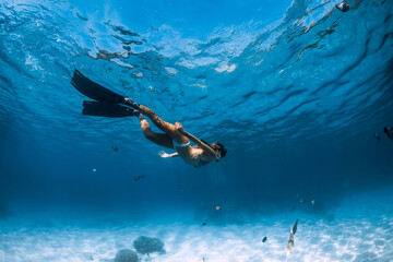 Woman freediver glides underwater with fins over sandy bottom.