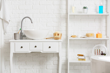 Table with modern sink and shelf unit with different bath supplies near white brick wall