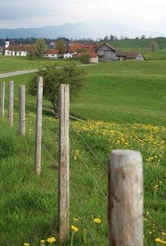 Weidezaun im hügeligen Voralpenland, im Hintergrund ein kleines Dorf und Berge, Allgäu, Bayern
