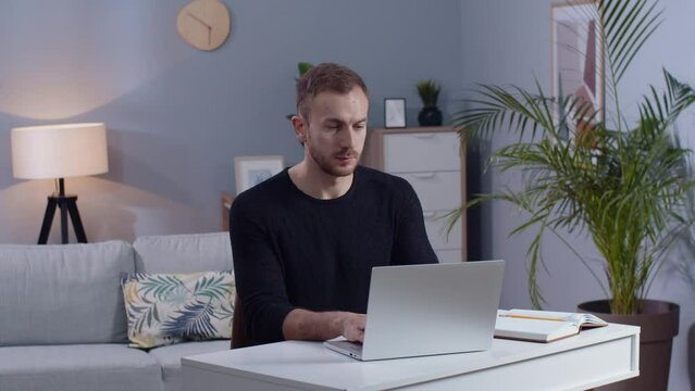 Shot of concentrated young man working on laptop in living room. Guy with beard touching aching neck, sitting at couch. Healthcare concept. Remote work, freelancer