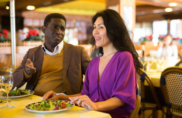 Fototapeta premium Cheerful african american man flirting with attractive asian woman during dinner in restaurant..