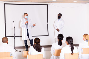 Portrait of confident successful male coach in face mask for disease prevention giving speech at medical conference