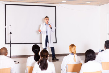 Confident male speaker in white coat giving presentation from stage at medical conference