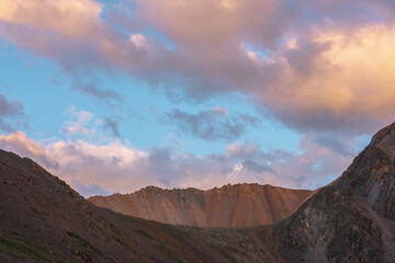 Obraz premium Dramatic landscape with sunlit wide sharp mountain ridge under clouds in sunset colors at changeable weather. Atmospheric mountain scenery with large sharp rocks on ridge top under sunset cloudy sky.