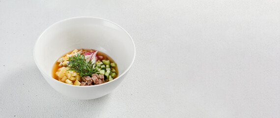Traditional russian soup - okroshka with fresh vegetables, meat and kvass. Cold summer soup on white table. Composition with traditional russian food and spices. Traditional soup with copy space.