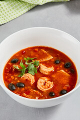 Fish tomato soup with shrimps and black olives. Seafood soup with prawns in white ceramic bowl on concrete background. Fish dish with seafood. Tomato soup in minimal composition.