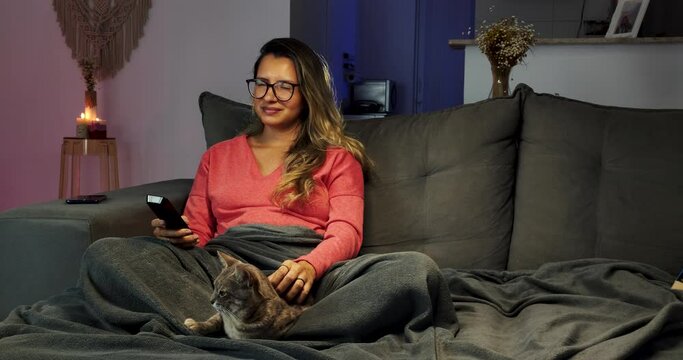 Jovem mulher sentada no sofá com um controle remoto na mão e acariciando um gato