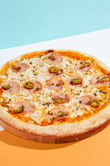Italian pizza with ham and jalapeno on coloured background. Spicy pizza with jalapeno in minimal style on orange and blue colours. American pizza delivery concept with color backdrop