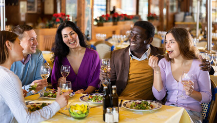 Multiracial group of happy adult people having fun in cozy restaurant during dinner, chatting and drinking wine..