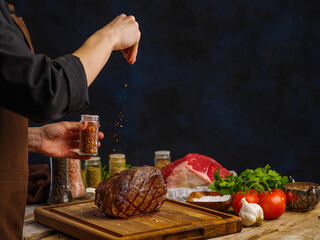 Fototapeta na wymiar Cooking ham on a wooden cutting board by the hands of a professional chef. The chef salts the baked ham. Vegetables, herbs, spices on a wooden table, dark blue background. Festive meat dish.